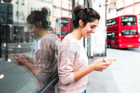 Téléchargez les photos : Femme heureuse à Londres - jeune femme caucasienne souriante utilisant son smartphone et s'appuyant contre la vitrine - style de vie et bonheur à Londres - en image libre de droit