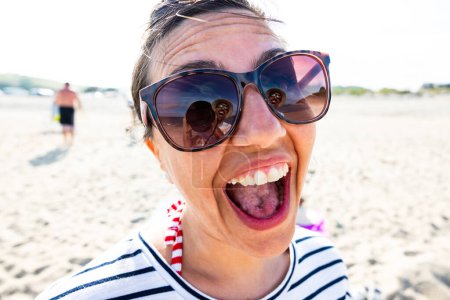 Foto de Primer plano de una mujer alegre en gafas de sol con una expresión lúdica en la playa y el marido fotógrafo reflexión en las gafas - Mujer alegre sobresaliendo lengua en el día de playa soleado - Imagen libre de derechos