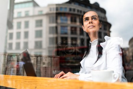 Foto de Mujer de negocios pensativa que trabaja desde un café en la ciudad - Mujer en la mesa con computadora y café, trabajando desde lejos durante un descanso - Estilo de vida de personas auténticas en Londres - Imagen libre de derechos