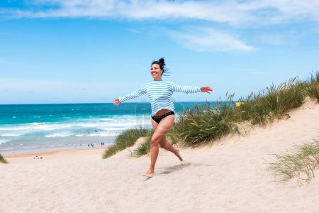 Téléchargez les photos : Joyeux jeune femme au bord de la mer en Cornouailles - Rire belle femme courant sur les dunes de sable avec l'océan en arrière-plan par une journée ensoleillée - Voyage et concepts de bonheur - en image libre de droit