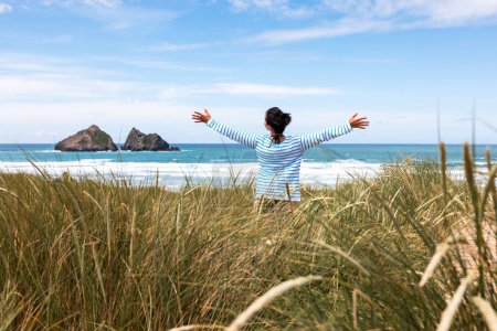 Téléchargez les photos : Femme au bord de la mer en Cornouailles regardant la mer et levant les bras - Vue arrière d'une femme debout sur des dunes de sable avec l'océan en arrière-plan par une journée ensoleillée - concepts de voyage et de bonheur - en image libre de droit