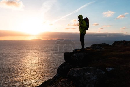 Téléchargez les photos : Explorateur admirant le coucher de soleil pittoresque de l'océan debout sur une falaise surplombant l'horizon - Silhouette de randonneur portant un sac à dos profitant de la solitude et de la réflexion dans la lueur du soir - en image libre de droit
