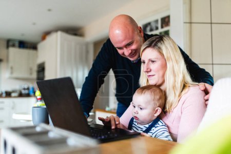Téléchargez les photos : Véritable prise de vue à domicile avec une jeune famille travaillant à domicile à l'aide d'un ordinateur et ayant un nouveau-né garçon dans le salon - Concepts du travail à distance, enfants, parentalité, mode de vie au Royaume-Uni - en image libre de droit