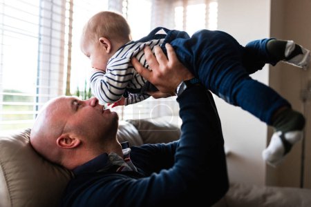 Téléchargez les photos : Heureux père et bébé garçon sur le canapé à la maison le tenant et profitant du temps ensemble - Style de vie et concepts de famille - en image libre de droit