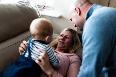 Téléchargez les photos : Authentic shot of happy family portrait with parents laughing and looking at their son while holding him on the sofa at home and enjoying time together - Lifestyle and family concepts - en image libre de droit