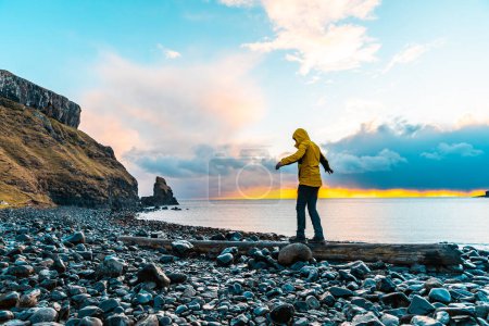 Téléchargez les photos : L'homme pendu dans la balance sur une bûche au bord de la mer en Écosse - Randonneur s'amuser à la belle destination baie de Talisker après la pluie - Filtre appliqué, concepts de voyage et d'aventure - en image libre de droit