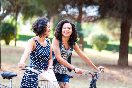Téléchargez les photos : Deux femmes souriantes profitant d'une journée ensoleillée à vélo ensemble à l'extérieur - Des amis joyeux sur une promenade à vélo dans le parc - en image libre de droit