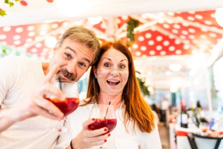 Téléchargez les photos : Joyeux couple d'âge moyen qui trinque avec des verres de vin sous une verrière à rayures rouges et blanches à Barcelone - Couple joyeux qui boit un verre dans un café extérieur à Barcelone - en image libre de droit