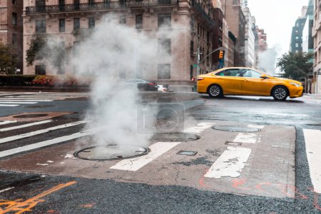 Téléchargez les photos : Une rue urbaine animée avec de la vapeur provenant d'un trou d'homme et un taxi en mouvement à New York Scène de rue torride avec taxi jaune dans la ville - en image libre de droit