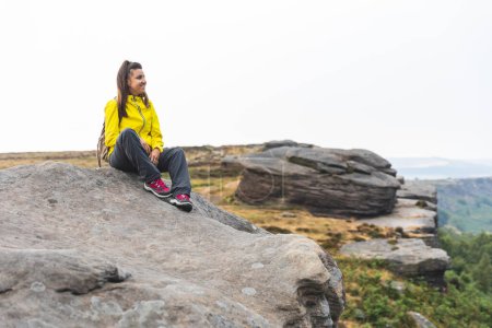 Téléchargez les photos : Jeune femme brune caucasienne reposant sur une falaise rocheuse et admirant la vue après une randonnée dans le Peak District - Femme assise au sommet d'une montagne - en image libre de droit