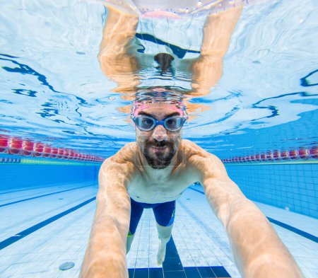 Foto de Primer plano de un alegre nadador masculino con gafas extendiéndose bajo el agua en una piscina clara - Vista submarina de un nadador sonriente en la piscina - Imagen libre de derechos