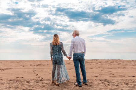 Back view of an elderly couple holding hands while walking on the sandy beach, serene sky above - Mature couple holding hands on beach at sunset