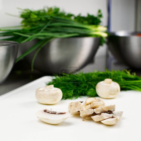 Téléchargez les photos : Fresh sliced mushrooms on a cutting board. In the background are green onions, parsley and dill in a stainless steel bowl - en image libre de droit