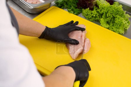 Téléchargez les photos : Hands of the cook in black gloves cut fresh chicken meat on a cutting board - en image libre de droit