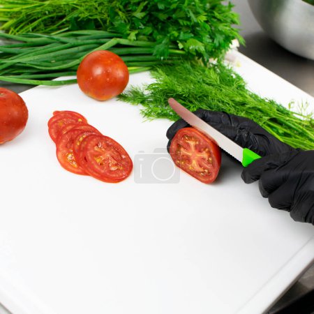 Foto de Las manos de cocinero en guantes de goma negro corta tomates. En la mesa hay perejil, eneldo y cebollas verdes.. - Imagen libre de derechos