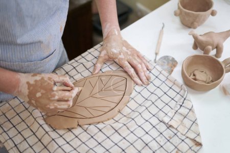 woman creates pattern on a leaf shaped piece of clay by hands in artistic studio.