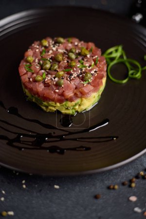 Tuna and avocado tartare with sesame seeds and capers on a dark ceramic plate.