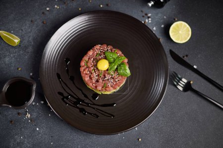 Tuna and avocado tartare with sesame seeds, capers and egg yolk on a dark ceramic plate.