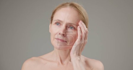 Anti-Age, Beauty, Gesundheit und Pflegekonzept für trockene Haut - schöne reife Kaukasierin mittleren Alters in ihren 50ern berührt ihre Gesichtshaut und blickt mit einem leichten Lächeln auf grauem Hintergrund in die Kamera
