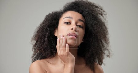 Schönheits- und Gesundheitskonzept - schöne Afroamerikanerin mit lockiger Afrofrisur und sauberer, gesunder Haut berührt ihre Wange und ihr Gesicht mit der Hand, posiert und blickt in die Kamera. Hoch