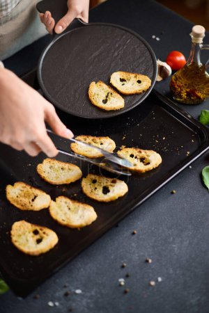 woman puts baguette chips from baking tray onto black ceramic serving board.