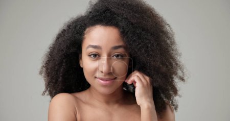 Concepto de belleza y salud: la hermosa mujer afroamericana con un peinado afro rizado y una piel limpia y saludable toca su mejilla y su cara con su mano, posando y mirando a la cámara. Alto.