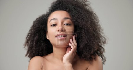 Concepto de belleza y salud: la hermosa mujer afroamericana con un peinado afro rizado y una piel limpia y saludable toca su mejilla y su cara con su mano, posando y mirando a la cámara. Alto.