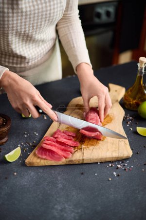Woman cut tuna steak into slices on a wooden cutting board at domestic kitchen.