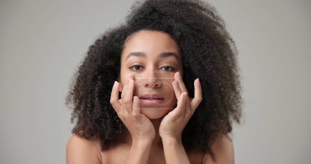 Concept beauté et soins de santé - belle femme afro-américaine avec une coiffure afro bouclée et une peau propre et saine touchant le visage avec la main, faisant un massage facial, posant et regardant la caméra. Haut