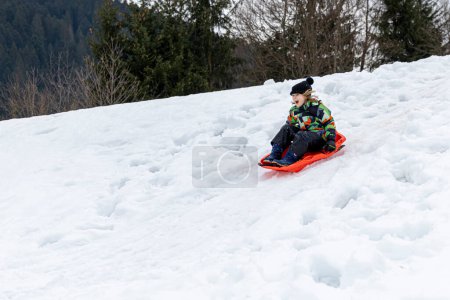 Téléchargez les photos : Little caucasian girl bobsled down a snowy slope. Amused expression - en image libre de droit