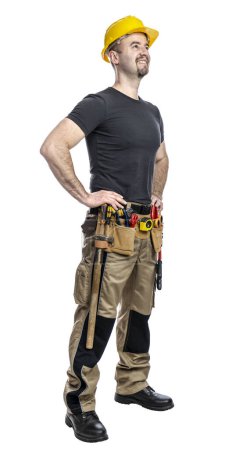 Téléchargez les photos : Travailleur de la construction confiant se tient avec les mains sur les hanches, portant un casque dur et une ceinture d'outils, isolé sur blanc - en image libre de droit