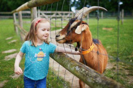 Foto de Adorable niñita alimentando cabra en la granja. Niña familiarizándose con los animales. Agricultura y jardinería para niños pequeños. Actividades al aire libre para niños - Imagen libre de derechos