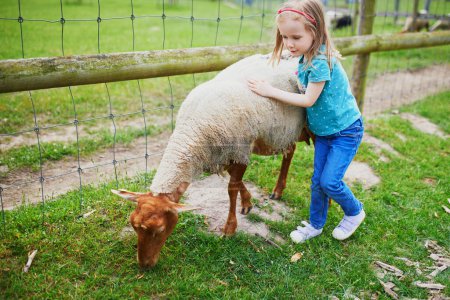 Foto de Adorable niña jugando con cabras y ovejas en la granja. Niña familiarizándose con los animales. Agricultura y jardinería para niños pequeños. Actividades al aire libre para niños - Imagen libre de derechos