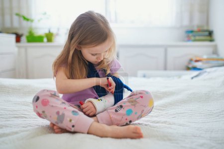 Photo for Adorable preschooler girl with a broken arm at home on the bed draws with felt-tip pens on an orthopedic cast. How to have fun with a broken limb. Broken arm in a cast in kids - Royalty Free Image
