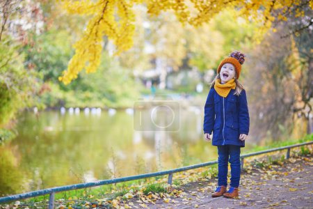 Téléchargez les photos : Adorable fille préscolaire profitant d'une belle journée d'automne ensoleillée en plein air. Joyeux enfant rassemblant des feuilles d'automne à Paris, France. Activités d'automne en plein air pour enfants - en image libre de droit