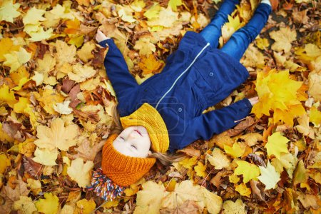 Téléchargez les photos : Adorable fille préscolaire profitant d'une belle journée d'automne ensoleillée en plein air. Joyeux enfant couché sur le sol couvert de feuilles d'automne à Paris, France. Activités d'automne en plein air pour enfants - en image libre de droit