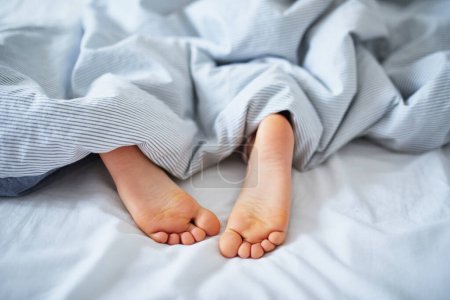 Photo for Little childs feet in bed covered with blanket. Kid sleeping in a comfortable bed. Early morning at home - Royalty Free Image
