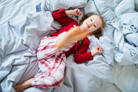 Photo for Happy preschooler girl wearing Christmas pajamas, playing on bed. Celebrating seasonal holidays with kids at home - Royalty Free Image