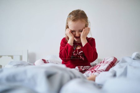 Photo for Unhappy preschooler girl wearing Christmas pajamas, sitting on bed. Celebrating seasonal holidays with kids at home - Royalty Free Image