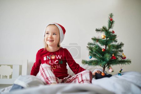 Photo for Happy preschooler girl wearing Christmas pajamas and playing with Christmas decorations on bed. Celebrating seasonal holidays with kids at home - Royalty Free Image