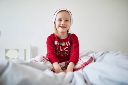 Photo for Happy preschooler girl wearing Christmas pajamas and red Santa hat, playing on bed. Celebrating seasonal holidays with kids at home - Royalty Free Image