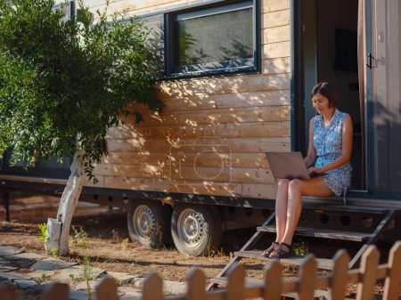 Glückliche junge Asiatin mit Laptop in der Nähe ihres winzigen Hauses, Wochenendausflug und abgelegener Büroidee. Tiny Houses und Small Living Konzept. Sommergarten