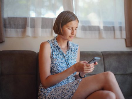 Photo for Relaxed young woman holding smartphone using mobile app, texting, searching for e-commerce offers on mobile device sitting on sofa at home. - Royalty Free Image