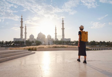 Foto de Viaje a los Emiratos Árabes Unidos, feliz joven viajera asiática con mochila y sombrero en Wahat Al Karama u Oasis de Dignidad, monumento permanente a sus mártires, y la Gran Mezquita Shaikh Zayed. - Imagen libre de derechos