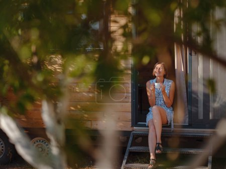 Photo for Woman is drinking coffee , morning routine near her Tiny house. First property. Small apartment in summer garden. Minimalism. Moving in. Living alone. Charming trailer house - Royalty Free Image