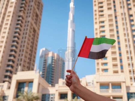 Primer plano de una mano femenina sosteniendo diminuta bandera de los Emiratos Árabes Unidos contra el horizonte de Dubai. Foto del espíritu de celebración del Día Nacional de los EAU.