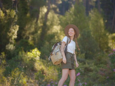 Caucasian ginger young beautiful female backpacker traveling alone in forest. Attractive traveler look around and explore while walk in nature wood with happiness and fun during holiday vacation trip.