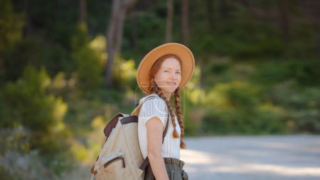Caucasian ginger young beautiful female backpacker traveling alone in forest. Attractive traveler look around and explore while walk in nature wood with happiness and fun during holiday vacation trip.
