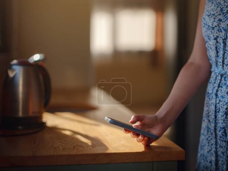 Photo for Female hand with smartphone in the kitchen, with morning sun rays. Tiny houses and Small Living concept. - Royalty Free Image