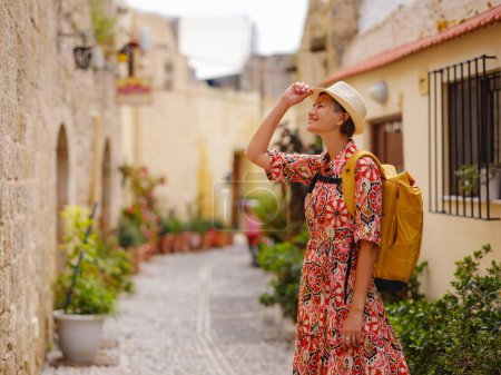 Young Asian woman in red dress and backpack walks and looks at cozy narrow streets of old city. Tourism, vacation, and discovery concept, female traveler visiting southern Europe, Rhodes island Greece